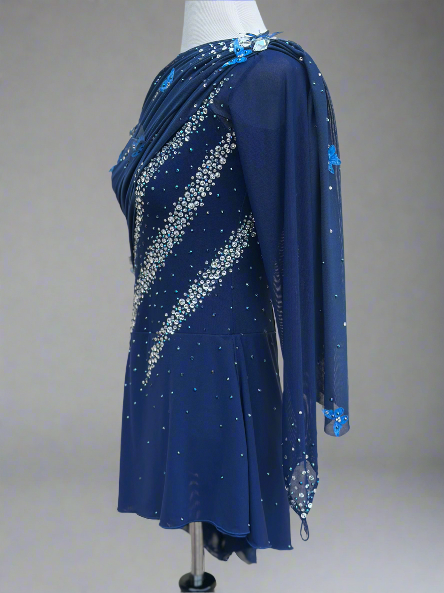 Elegant butterfly figure skating dress in deep blue with crystals, draped design, and open back – sparkle on ice.