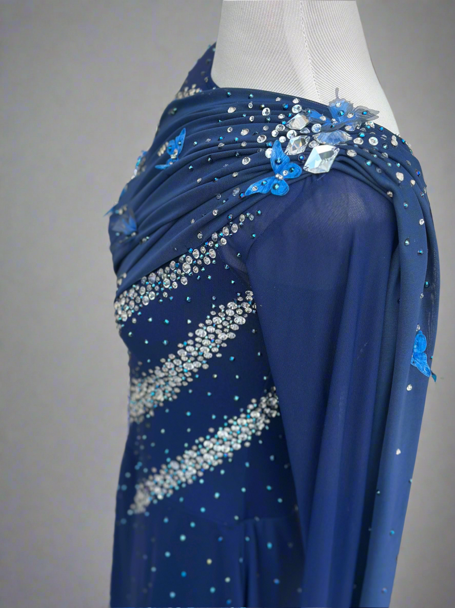 Elegant butterfly figure skating dress in deep blue with crystals, draped design, and open back – sparkle on ice.